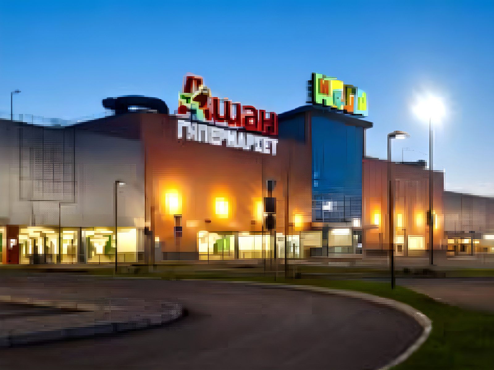 Shopping-centre frontage at dusk: a lit colonnade of shopfronts under the MEGA roof signage and a curving access road in front