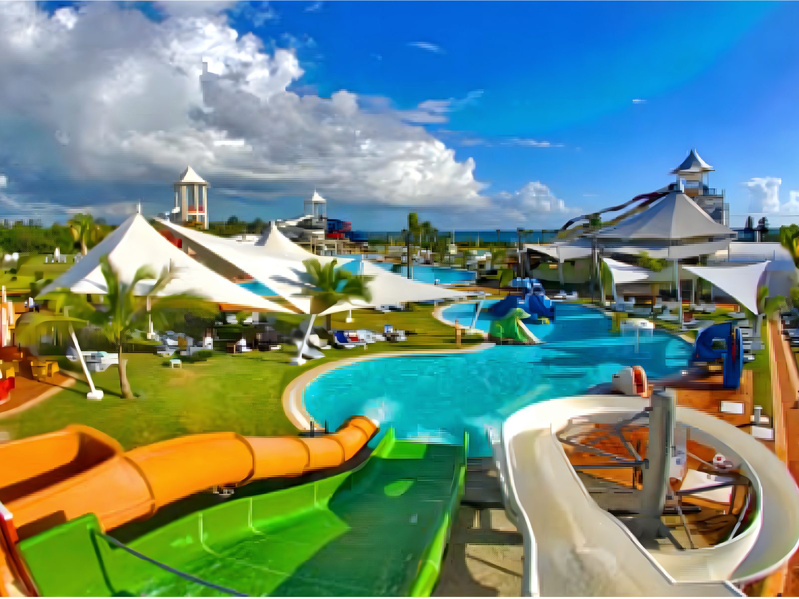 Aerial view over the park's slide tower: white shade sails, a turquoise lagoon pool, an orange flume and a green racer slide, the sea beyond