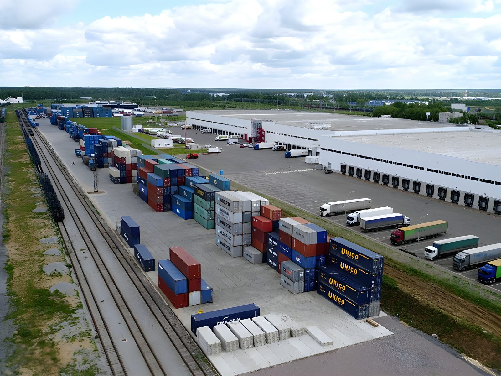 Aerial view of the logistics yard: stacked containers beside rail sidings and a long warehouse with a full line of loading docks